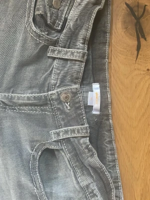 Grå jeans från Gina Tricot med detaljer - Snygga grå jeans från Gina med markerade sömmar och coola fickdetaljer både fram och bak. Jeansen har knappar på fickorna och breda hällor för bälte. Perfekta för dig som gillar en avslappnad men trendig stil.