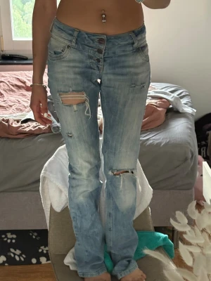 Ljusblå slitna bootcut jeans - Säljer ett par ljusblå bootcut jeans med slitningar och hål på låret och knät. Jeansen har knappgylf, fem fickor och en avslappnad passform. Perfekta för en avslappnad och trendig look.