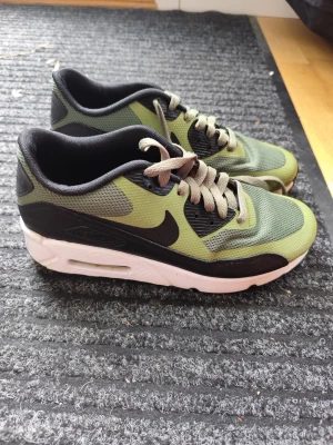 Nike Air Max gröna sneakers - Nike Air Max sneakers i grönt mesh med svarta och vita detaljer. Skorna har en klassisk låg siluett, vit sula med synlig Air-enhet och beige skosnören. Perfekta för dig som gillar streetwear och sportig stil.