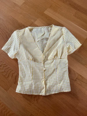 Gulvitrutig blus med v-ringning - Söt blus i gulvitt rutigt mönster med djup v-ringning och knappar framtill. Modellen har korta ärmar och figurnära passform. Perfekt till jeans eller kjol för en fräsch sommarlook. Materialet känns lätt och luftigt.