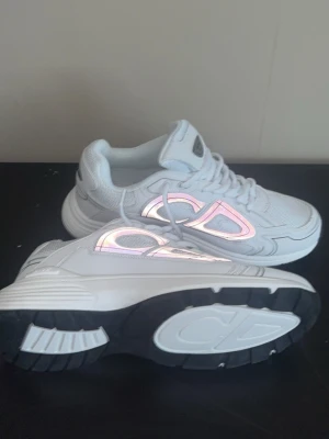 Vita Dior B22 sneakers med reflexdetaljer - Snygga vita Dior B22 sneakers med reflekterande CD-logga på sidorna. Skorna har en chunky sula i vitt och svart, mesh och läderdetaljer samt klassisk snörning. Perfekta för dig som vill sticka ut med exklusiva och trendiga sneakers.