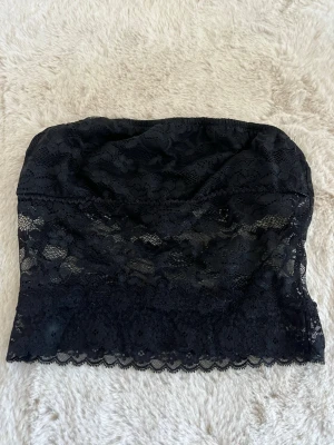 Svart bandeau tubtopp i spets - Säljer en svart bandeau topp i spets med fina blommiga detaljer. Toppen är axelbandslös och har en mjuk, stretchig passform. Perfekt att styla under en skjorta eller kavaj för en trendig look.