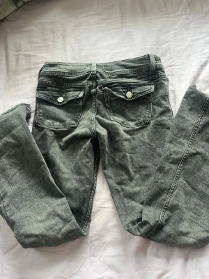 Gråa bootcut jeans med fickdetaljer - Säljer ett par gråa bootcut jeans med dubbla knappar fram och snygga fickor med lock och knapp bak. Jeansen har en klassisk femficksmodell och är tillverkade i stretchigt denimtyg för skön passform.