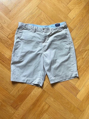 Beige shorts från Polo Ralph Lauren - Klassiska beige shorts från Polo Ralph Lauren med raka ben, bälteshällor och sidofickor. Baksidan har två fickor med knappstängning. Perfekta för sommaren och tillverkade i mjuk bomull för en skön känsla.