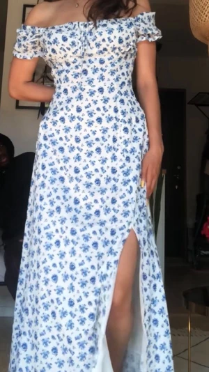 Blommig maxi klänning - Första bilden är när jag har på mig den. Långklänning med blått blommönster och vit. Klänningen har puffiga kortärmar, fyrkantig ringning/ off shoulder och en  knytning framtill. 