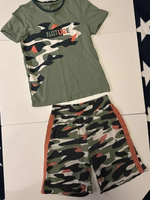 Grönt Kamouflage set strl 116/122 med t-shirt och shorts - Grönt kamouflage set med t-shirt och shorts. Grönt set strl 116/122 camouflage shorts och tshirt   Byxan har några små hål nedertill - se bild  Ett trådsläpp  Bonprix Sommarset  Byxor  Tröja  T-shirt  Militär Sommar