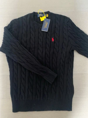 Svart kabelstickad tröja Polo Ralph Lauren - Kabelstickad svart tröja från Polo Ralph Lauren med rund halsringning och röd broderad logga på bröstet. Tröjan har ribbade muddar vid ärmslut och nederkant. Perfekt för dig som gillar klassisk och stilren design.