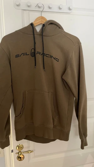 Olivgrön hoodie från Sail Racing - Olivgrön hoodie från Sail Racing med svart logga tryckt på bröstet och en mindre logga på huvan. Klassisk känguruficka framtill och svarta dragsnören i huvan. Tillverkad i mjukt bomullsmaterial, perfekt för en avslappnad stil.