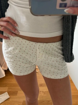 Brandy Melville shorts - Söta vita pyjamasshorts med små rosa blommor och ribbad struktur. Shortsen har knappgylf framtill och är gjorda i ett mjukt, stretchigt material som passar perfekt för mysiga hemmakvällar.