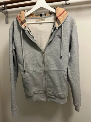 Grå zip hoodie från Burberry - Snygg grå zip hoodie från Burberry med klassiskt rutigt foder i huvan och dragsko. Hoodien har två fickor framtill och diskret Burberry-logga på bröstet. Tillverkad i mjukt material som känns skönt mot huden. Perfekt för en avslappnad och trendig look.