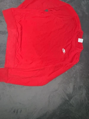 Röd stickad tröja från Nike - Säljer en röd stickad tröja från Nike med klassisk rund halsringning och diskret Nike-logga broderad i vitt på bröstet. Tröjan har långa ärmar och ribbade muddar vid ärmslut och nederkant. Perfekt för dig som gillar sportig och enkel stil.