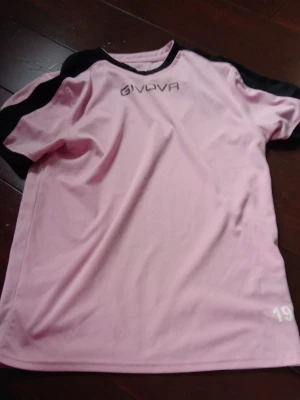Rosa sporttröja från Givova - Säljer en rosa sporttröja från Givova med svarta detaljer på axlar och krage. Tröjan har korta ärmar och är tillverkad i ett lätt och snabbtorkande material, perfekt för träning eller match. Nummer 19 är tryckt i vitt nere på framsidan.