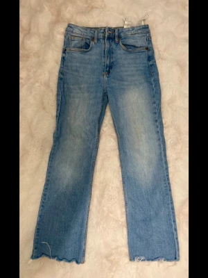 Ljusblå raka jeans med rå kant - Säljer ett par ljusblå jeans med rak passform och trendigt råa, fransiga benslut. Jeansen har klassisk femficksdesign och är tillverkade i denim. Perfekta för en avslappnad och cool stil.
