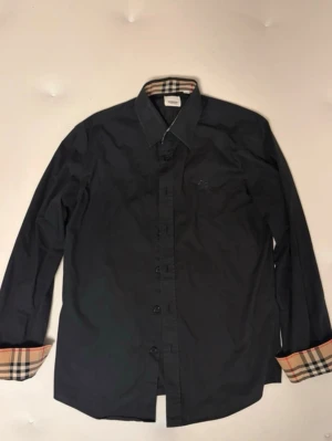Burberry skjorta  - Stilren svart skjorta från Burberry med klassiskt rutigt mönster på insidan av kragen och manschetterna. Skjortan har knappar framtill och lång ärm. Perfekt för dig som gillar exklusiva detaljer och tidlös design.