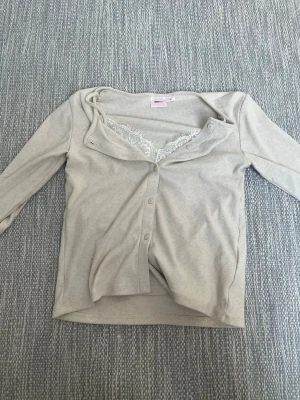 Beige ribbad kofta från Gina Tricot - Säljer en beige ribbad kofta från Gina Tricot med knappar framtill och spetsdetalj vid halsringningen. Koftan har långa ärmar och en mjuk, stretchig känsla.