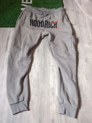 Gråa mjukisbyxor från Hoodrich - Snygga gråa mjukisbyxor från Hoodrich med svart och orange logga framtill. Byxorna har dragsko i midjan, muddar vid bensluten och en praktisk ficka på sidan. Tillverkade i mjukt och bekvämt material, perfekta för chill dagar.