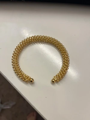 Guldigt pärlarmband i spiraldesign - Snyggt armband i guldfärg med små pärlor i spiralformad design. Armbandet är stelt och öppet i ändarna med två guldfärgade kulor som avslut. Perfekt accessoar för att lyfta din outfit med en touch av bling.