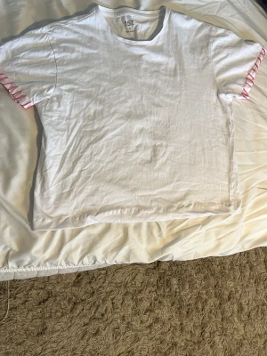 Vit t-shirt med rosa snörning - Vit t-shirt i bomull med korta ärmar och unik rosa snörningsdetalj längs ärmsluten. Enkel och clean design med en twist som gör den extra intressant. Perfekt för dig som gillar att sticka ut lite utan att det blir för mycket. Har gjort den rosa snörningen själv, och dessutom sytt upp tröjan så att den blev lite kortare i längden!!💓💓