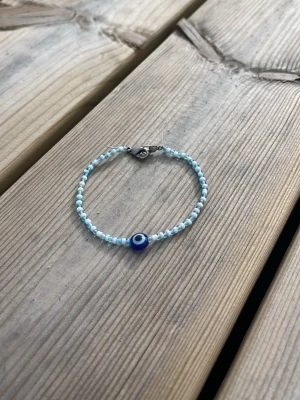 Pärlarmband med blått öga - Snyggt handgjort armband med små vita och ljusblå pärlor samt en större blå pärla med ögonmotiv i mitten. Armbandet har ett metallspänne och passar perfekt för dig som gillar trendiga accessoarer med en unik touch. Armbandet är 17,5 cm i längden.