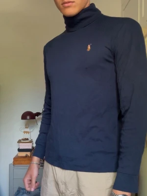 Mörkblå polotröja från Ralph Lauren - Mörkblå långärmad polotröja från Ralph Lauren med klassisk broderad logga på bröstet. Tröjan är i mjukt bomullsmaterial och har en tight passform med hög krage. Perfekt för en stilren och enkel look. (Passform: Jag på bilden är 190 lång och väger 80kg)
