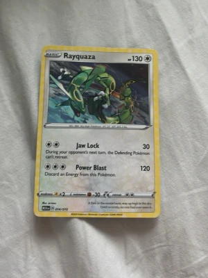 Rayquaza Pokémonkort - Säljer ett Rayquaza Pokémonkort med gul ram och illustration av Rayquaza i grönt och gult. Kortet har attackerna Jaw Lock och Power Blast samt 130 HP. Text och symboler i svart och silver. Perfekt för samlare eller Pokémonfans.