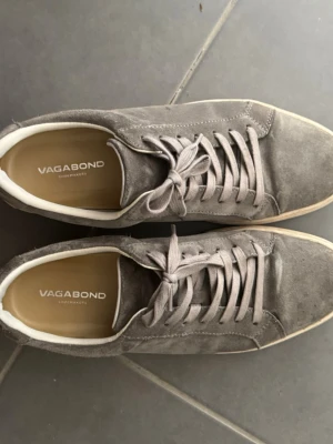 Gråa Vagabond sneakers i mocka - Snygga grå sneakers från Vagabond i mjuk mocka med vita sulor och matchande grå skosnören. Klassisk låg modell som passar perfekt till jeans eller chinos. Diskret logga på insidan och stilren design för en clean look.
