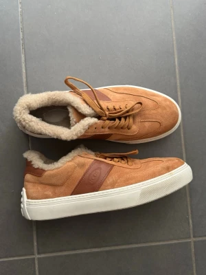 Tod’s sneakers – Italienska premiumsneakers | Mycket fint skick - Snygga bruna sneakers från TOD'S i mocka med vit sula och mjukt foder i fuskpäls. Skorna har bruna läderdetaljer och klassisk snörning. Perfekta för dig som vill ha både stil och komfort under kallare dagar.