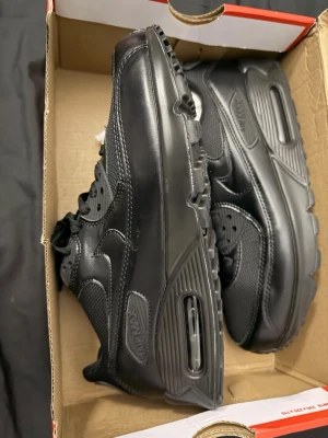 Nike Air Max 90 helsvarta sneakers - Snygga helsvarta Nike Air Max 90 sneakers med klassisk siluett. Skorna har ovandel i en mix av läder och mesh, samt den ikoniska Air Max-dämpningen i sulan. Perfekta för dig som gillar stilrena och tidlösa sneakers med streetkänsla.