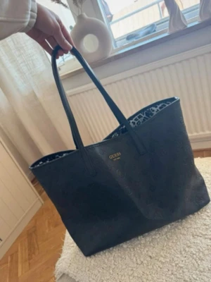 Svart shoppingväska från Guess - Snygg svart shoppingväska från Guess med präglat logomönster och guldfärgad logga framtill. Väskan har långa handtag och insidan är fodrad med ett djurmönstrat tyg. Perfekt för dig som vill ha plats för allt du behöver under dagen.