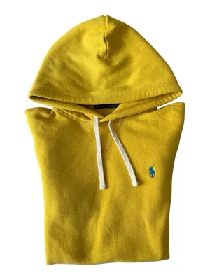 Gul hoodie från Polo Ralph Lauren - Säljer en snygg gul hoodie från Polo Ralph Lauren med vit dragsko och klassisk blå logga på bröstet. Hoodien har en stor huva, långärmad design och en praktisk magficka. Materialet är mjukt och bekvämt, perfekt för att ge din outfit en färgboost.