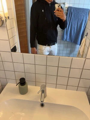 Svart hoodie från Polo Ralph Lauren - Säljer en svart hoodie från Polo Ralph Lauren med dragkedja framtill och klassisk huva. Diskret logga broderad på bröstet. Hoodien har fickor på sidorna och är tillverkad i mjukt material som känns skönt mot huden. Perfekt för en avslappnad och stilren look.