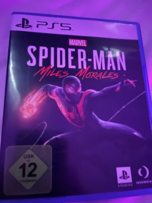 Spider-Man Miles Morales PS5 spel - Säljer Marvel's Spider-Man: Miles Morales till PS5. Spelet kommer i ett blått plastfodral med snyggt omslag och skiva med röd spindel på svart bakgrund. Perfekt för dig som gillar action och superhjältar!