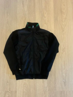 Svart vindjacka från Hugo Boss - Snygg svart vindjacka från Hugo Boss med broderad logga på bröstet i svart och grönt. Jackan har dragkedja framtill, hög krage med grön detalj på insidan och ribbade muddar vid ärmslut och nederkant. Perfekt för dig som vill ha en stilren och sportig look.