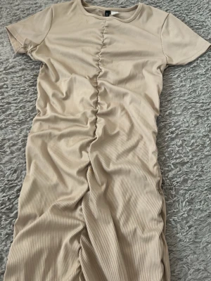 Beige ribbad klänning med snörning - Trendig beige ribbad klänning med korta ärmar och snygg snörning längs mitten framtill. Klänningen har en kroppsnära passform och är tillverkad i ett mjukt material som känns skönt mot huden. Perfekt för dig som gillar stilrena och moderna plagg.