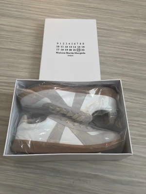 Maison Margiela vita sneakers - Maison Margiela sneakers i vitt läder med ljusgrå mockadetaljer och beige snören. Klassisk siluett med gummisula i brunt och diskret branding. Perfekta för dig som gillar minimalistisk och stilren design.