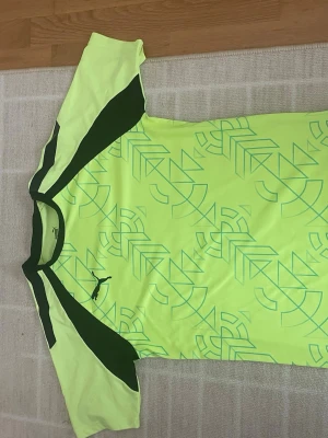 Neongul sporttröja från Puma - Snygg neongul sporttröja från Puma med mörkgröna detaljer på axlar och sidor. Tröjan har ett grafiskt mönster framtill och är tillverkad i ett lätt, snabbtorkande material som passar perfekt till träning eller match.
