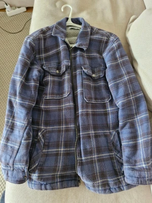 Blå-rutig overshirt med teddyfoder - Säljer en blå och grå-rutig overshirt med dragkedja och två bröstfickor med knappar. Insidan är fodrad med mjukt grått teddyfoder som gör den extra varm och skön. Perfekt lager-på-lager-plagg för kyliga dagar.