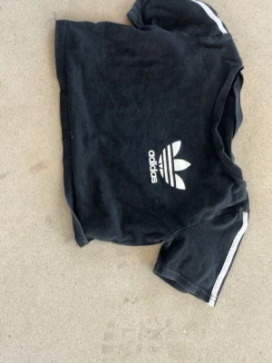 Svart Adidas t-shirt med logga - Cool svart mag  t-shirt från Adidas med den klassiska treklöver-loggan i vitt på bröstet och vita ränder på axlarna. T-shirten har korta ärmar och en avslappnad passform, perfekt för en sportig look.