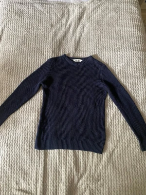 Mörkblå stickad tröja från H&M - Säljer en mörkblå stickad tröja från H&M med rund halsringning och lång ärm. Tröjan har en diskret strukturstickning och ribbade muddar vid ärmslut och nederkant. Perfekt basplagg för lager på lager och enkel att matcha.