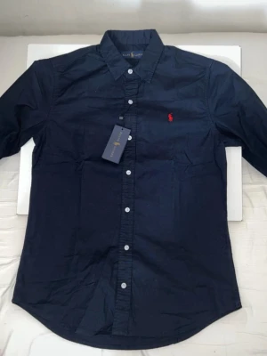 Mörkblå skjorta från Ralph Lauren - Klassisk mörkblå skjorta från Ralph Lauren med vit knappstängning och röd broderad logga på bröstet. Skjortan har lång ärm, krage och är tillverkad i bomull. Perfekt för en stilren och tidlös look.