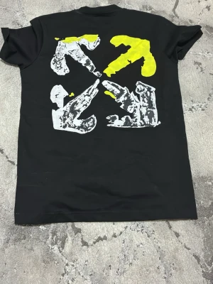 Svart Off-White t-shirt med tryck - Säljer en svart t-shirt från Off-White med coolt grafiskt tryck i gult och vitt både fram och bak. T-shirten har rund hals och korta ärmar. Perfekt för dig som gillar streetwear och vill sticka ut med en unik design.