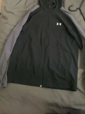Svart vindjacka från Under Armour - Snygg svart vindjacka från Under Armour med gråa ärmar och dragkedja framtill. Jackan har en vit logga på bröstet och är tillverkad i ett lätt och sportigt material, perfekt för aktiva dagar utomhus.