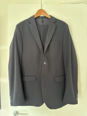 Marinblå kostym från Selected Homme - Snygg marinblå kostym från Selected Homme i slim fit-modell. Klassisk design med två fickor framtill, bröstficka och enkla knappar vid ärmslut. Perfekt för dig som vill ha en stilren och modern look. Använd ~3 gånger. Byxorna är i storlek 52, se mått på bilder för kavajen.