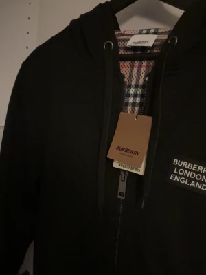 Svart Burberry zip hoodie - Snygg svart hoodie från Burberry med dragkedja framtill och klassisk logga 'BURBERRY LONDON ENGLAND' på bröstet. Hoodien har huva med dragsko och ett rutigt innerfoder som ger en exklusiv touch. Perfekt för dig som gillar stilrena och lyxiga plagg.