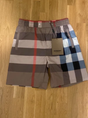 Rutig Burberry shorts  - Snygga shorts från Burberry med klassiskt rutigt mönster i brunt, beige, svart, vitt och rött. Modellen har elastisk midja och sidofickor. Perfekta för en trendig och avslappnad stil.