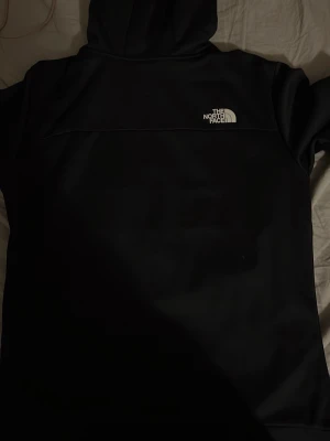 North face Hoodie - Snygg svart hoodie från The North Face med dragkedja framtill och stor logga på bröstet. Hoodien har huva och är tillverkad i ett mjukt och bekvämt material, perfekt för en avslappnad streetstyle-look. Knappt använd och har inga deffekter