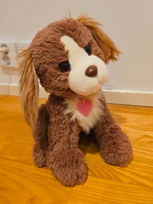 Brunt mjukisdjur hund med halsband - Mjukisdjur i form av en hund med brun och vit päls, stora svarta ögon och en rosa nos. Hunden har ett rosa halsband med blå hjärtan och ett rosa hjärthänge. Tillverkad i mjukt plyschmaterial och perfekt som gosedjur eller dekoration.
