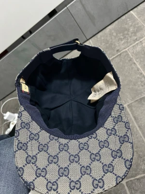 Gucci beige keps med blå GG-mönster - Säljer en beige keps från Gucci med klassiskt blått GG-monogram över hela kepsen. Kepsen har böjd skärm, justerbart band bak och en snygg detalj med blå och röd rand på sidan. Materialet är canvas med insida i mörk textil. Perfekt för dig som gillar exklusiva accessoarer.