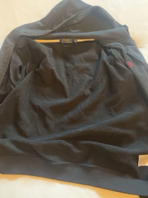 Svart hoodie från Polo Ralph Lauren - Svart hoodie från Polo Ralph Lauren med klassisk röd broderad logga på bröstet. Tröjan har huva, känguruficka och ribbade muddar. Tillverkad i mjukt bomullsmaterial som är skönt att ha på sig.