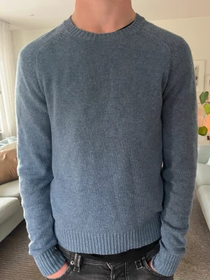 100% Ulltröja från Uniqlo - Mysig blå stickad tröja med rund hals och ribbade muddar vid ärmslut och nederkant. Tröjan har en enkel och stilren design, perfekt för lager-på-lager. Materialet är mjukt och ger en avslappnad känsla. Ett minimalt hål finns på bild nr 2.